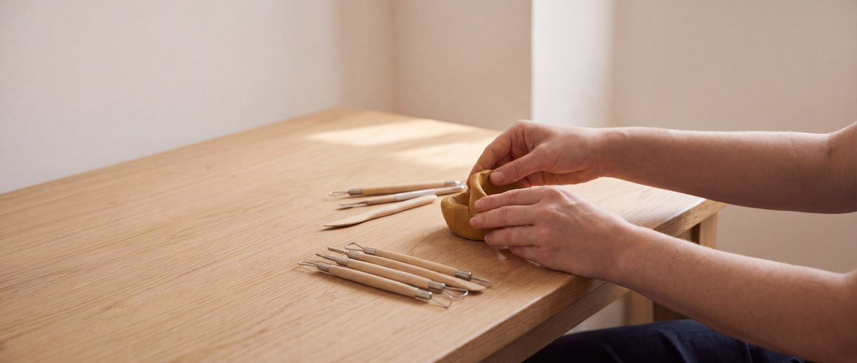 Handen vormen een kommetje van klei op een houten tafel, met boetseergereedschap ernaast