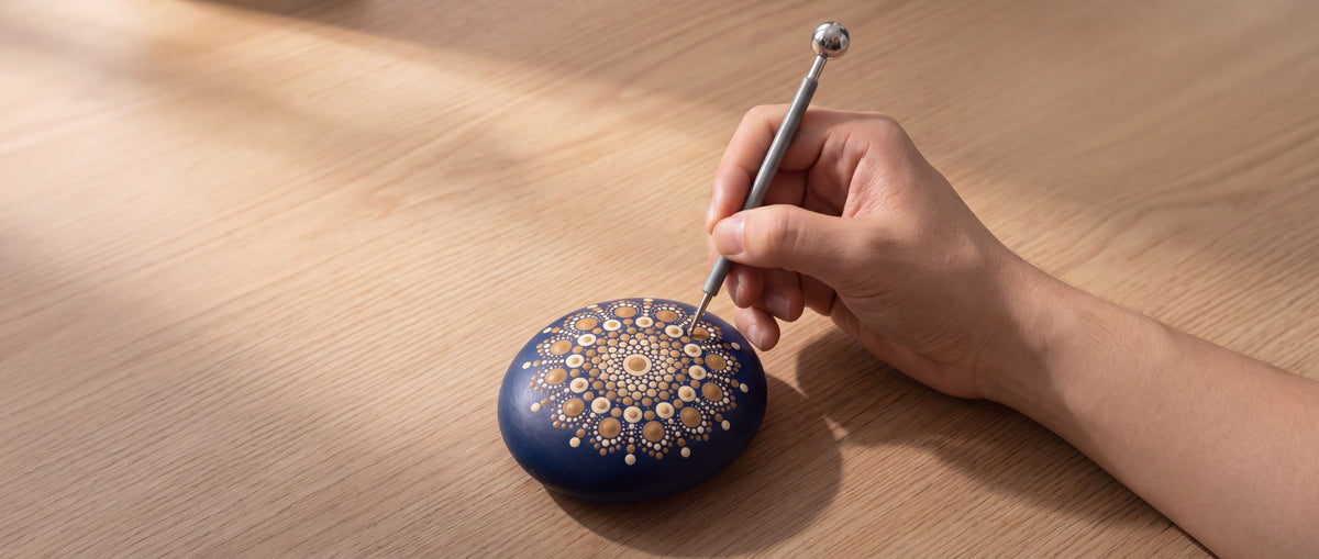 Een hand zet met een dotting tool stippen op een blauwe mandalasteen